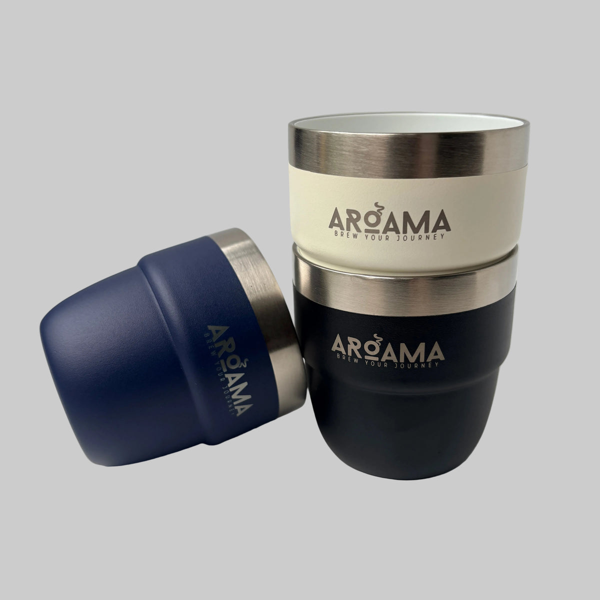 Three Arcama travel tumblers with stainless steel rims, shown in navy blue, cream, and black, with one tumbler tipped over on its side to display the lid.