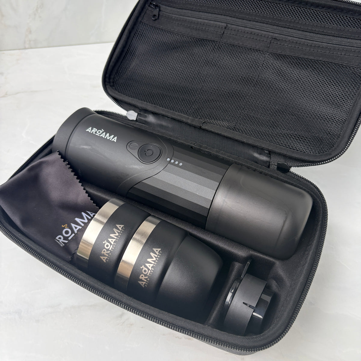A black Aroama portable espresso maker set displayed inside its open zippered carrying case, including the main device, a travel tumbler, and a small accessory, on a light marble surface.
