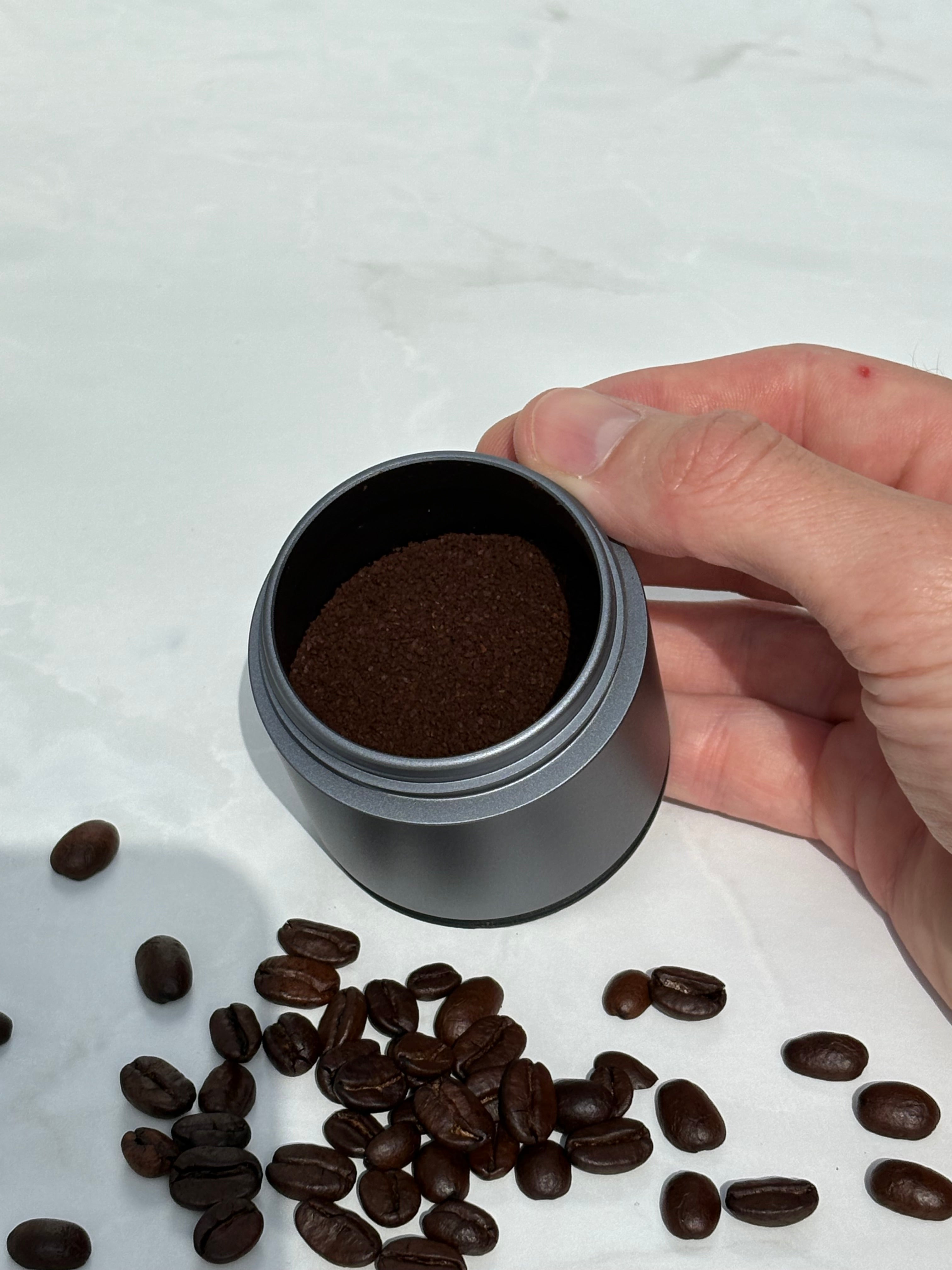 Hand holding a small container of ground coffee with scattered coffee beans on a light background