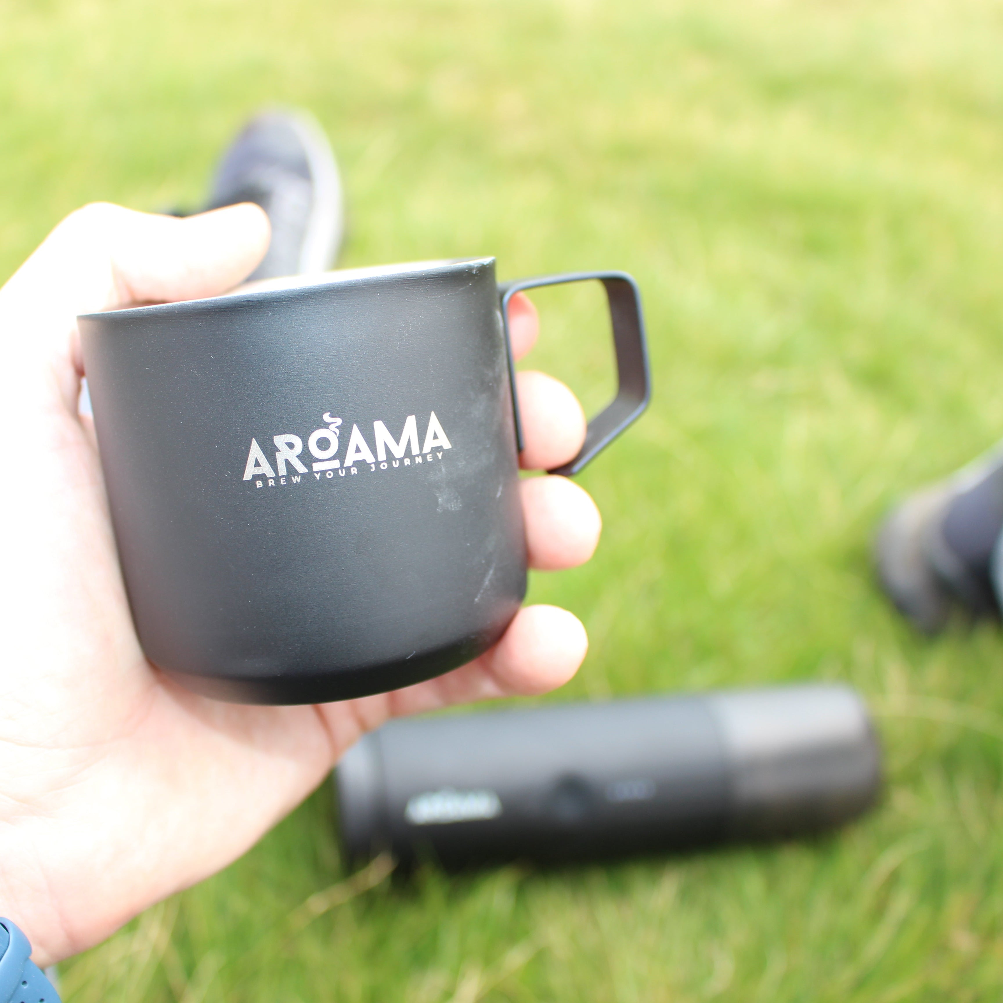 Person holding a black mug with 'AROMA' branding on grass