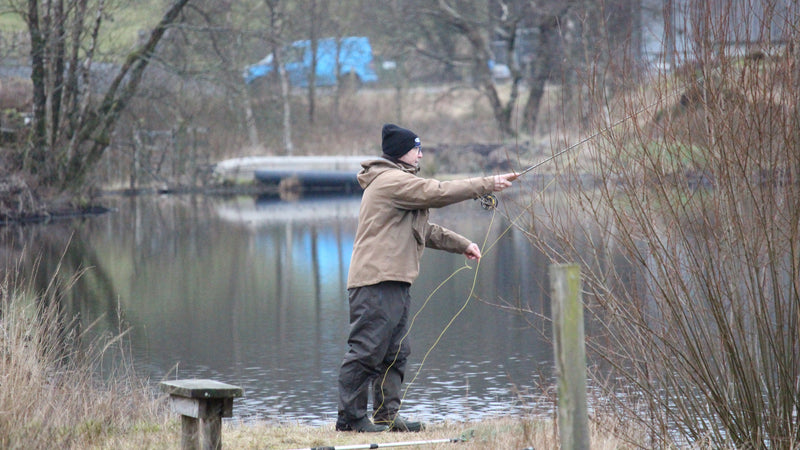 A man fly fishing on a calm lake, casting a rod with a line. He is wearing a tan jacket, dark trousers, and a black beanie.