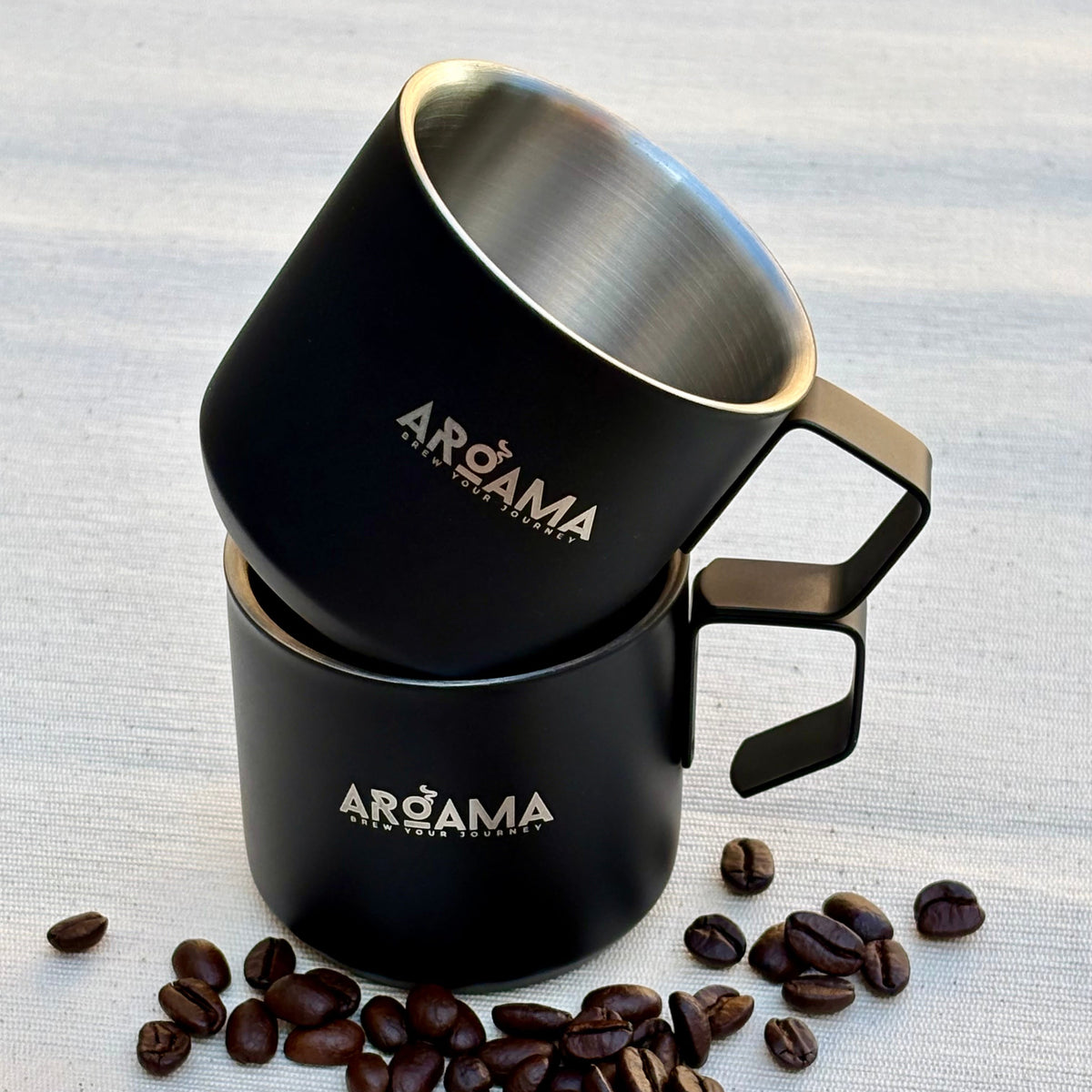 Two black mugs with 'AR0AMA' branding on a light gray surface with scattered coffee beans.