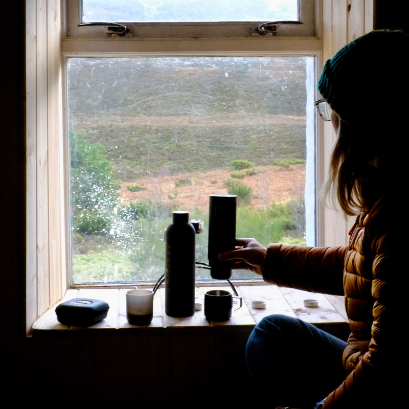 A person in a dark beanie and puffer jacket sits by a window, holding a black cylindrical mug or tumbler next to a tall black bottle and small cups on a windowsill, with an outdoor landscape visible behind the glass.